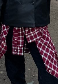 Personne portant un pantalon noir et un manteau noir avec une chemise à carreaux rouge et blanche nouée autour de la taille en plein air.