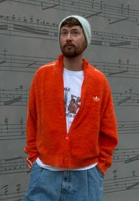 Orange fuzzy zip-up jacket with a high collar, worn over a white graphic tee. Paired with light blue denim shorts. Light grey beanie.