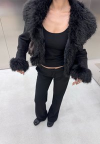 Sort faux fur jakke med stor krave, parret med en sort tætsiddende top og sorte bukser. Det overordnede look er monokromt og tekstureret.
