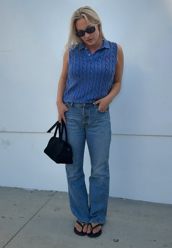 Vrouw met lang blond haar, zonnebril, blauwe mouwloze trui, jeans, zwarte sandalen, draagt een zwarte handtas, staand tegen een witte muur.