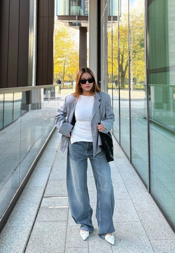 Blazer oversized gris claro, camisa blanca ajustada, vaqueros anchos azules y tacones blancos de punta; un bolso negro y gafas de sol completan el look.
