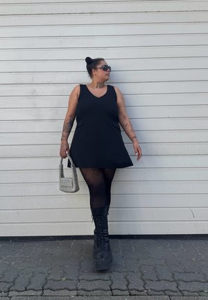 Vestido negro sin mangas con escote en V y bolsillos laterales, combinado con un bolso de mano negro texturizado y botas de plataforma de cordones hasta la rodilla.