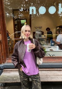 Jachetă bomber maro, tricou violet, pantaloni cu imprimeu leopard. Ținând o cană de cafea. Fundalul prezintă fereastra unui cafenea cu oameni în interior.