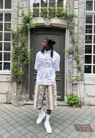 Camisa blanca con texto negro, pantalones cortos de estampado de serpiente, zapatillas blancas y una boina negra. Entorno al aire libre con follaje verde y una puerta gris.