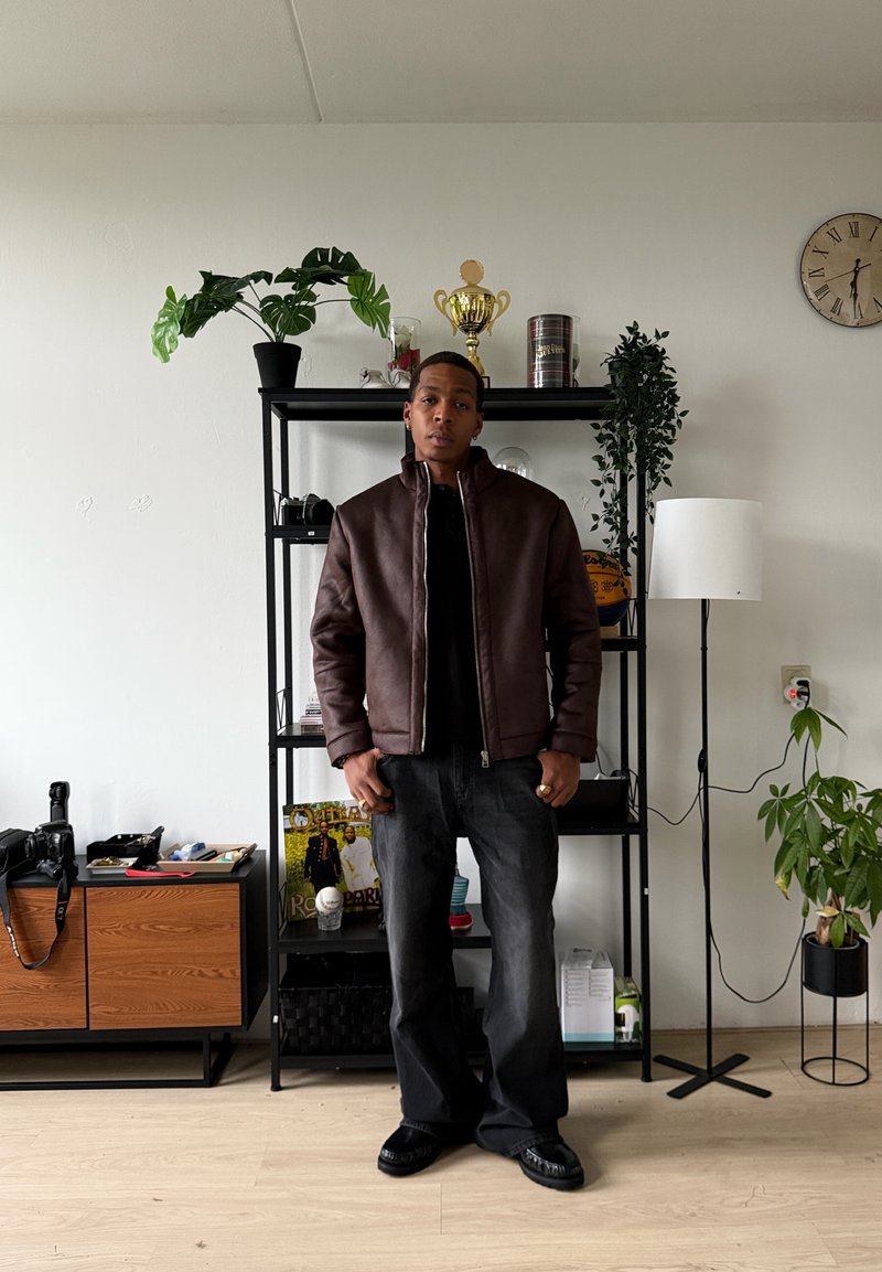 Veste aviateur en cuir marron avec un col montant, chemise noire, jean décontracté foncé et chaussures noires. Cadre intérieur avec étagères et plantes.