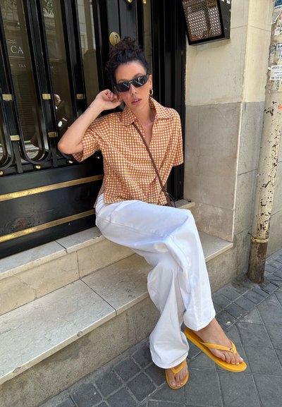 Modelo que lleva una camisa de cuadros amarilla y naranja, pantalones anchos blancos y chanclas amarillas. Sentada en un escalón de piedra contra una puerta negra.