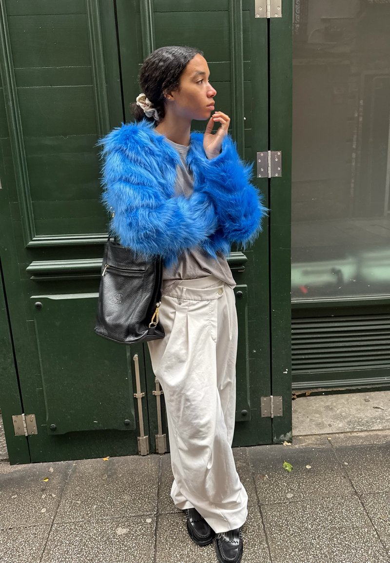 Jeune personne portant une veste en fausse fourrure bleue, un pantalon blanc et des chaussures noires, debout avec un sac à bandoulière noir contre une porte vert foncé.