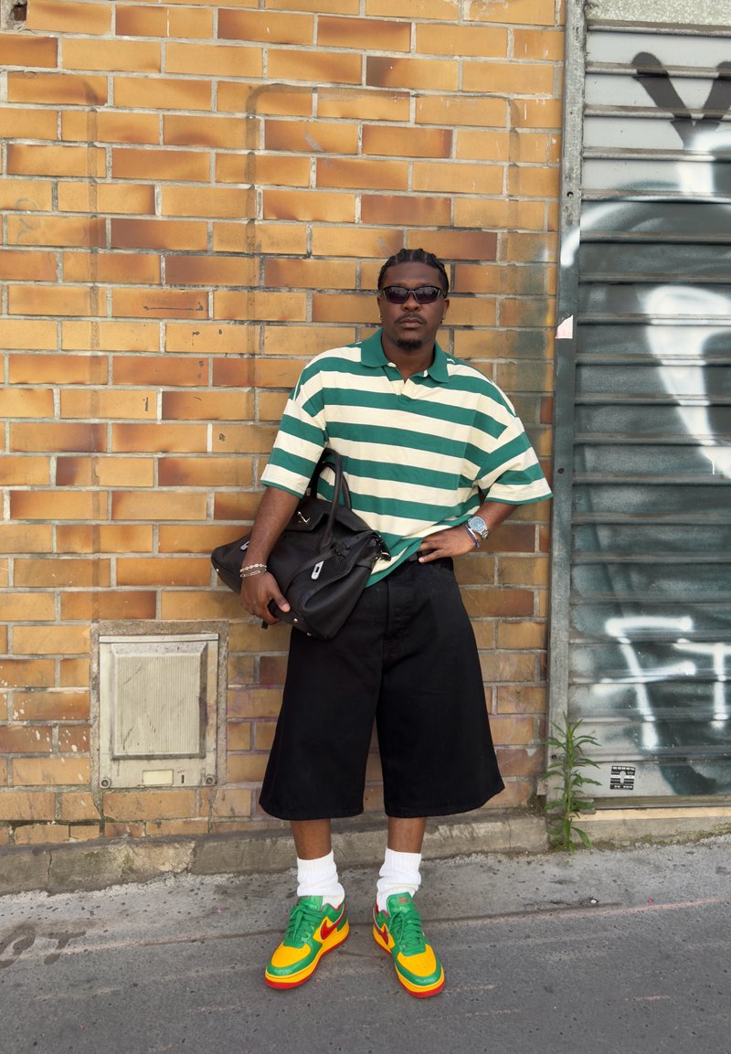 Homme portant une chemise rayée verte et blanche, un short noir, des baskets colorées et des lunettes de soleil, debout près d'un mur en briques avec un volet couvert de graffiti.