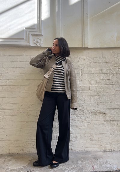 Chaqueta de cuero beige, suéter de cuello alto a rayas blanco y negro, pantalones anchos negros y zapatos negros. De pie contra una pared crema texturizada.