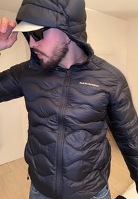 Άνδρας που φοράει μαύρο μπουφάν Peak Performance με κουκούλα, μαύρα γυαλιά ηλίου και λευκό καπέλο, κρατώντας το γείσο του καπέλου σε εσωτερικό χώρο.