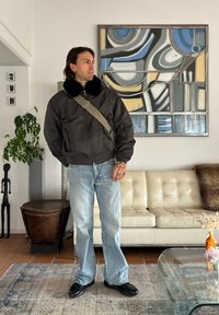 Veste bombardier marron avec col en fourrure, jean bleu clair, chaussures noires, debout dans un intérieur moderne avec une œuvre d'art abstraite et des plantes.