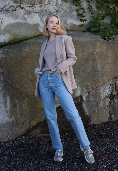 Mujer con blazer beige, suéter de punto, guantes, vaqueros azules y zapatillas deportivas de pie junto a una pared rocosa cubierta de hiedra trepadora.