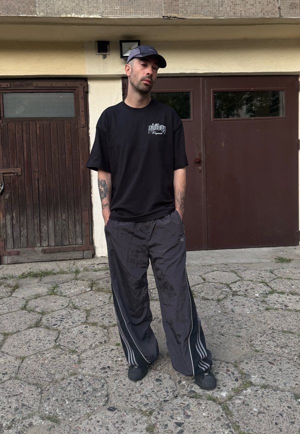 Homme avec des tatouages portant une casquette noire, un t-shirt noir et un pantalon noir ample, debout sur un pavé fissuré devant des portes en bois marron et en métal.