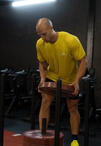 Moški z izpraznjeno glavo v svetlo rumeni majici dviguje okroglo rdečo utežno ploščo v telovadnici z vadbeno opremo v ozadju.