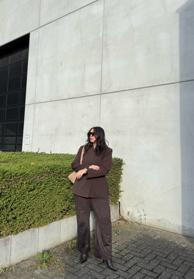 Chaqueta ajustada marrón y pantalones de pierna ancha a juego, combinados con botas de tacón negras. Los accesorios incluyen un bolso cruzado beige y gafas de sol.