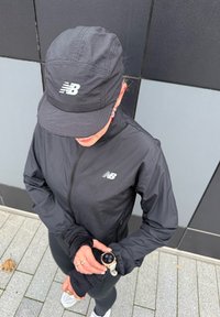Veste légère noire avec fermeture éclair frontale, arborant un logo sur la poitrine, associée à une casquette assortie et des leggings ajustés. Une montre est portée au poignet.