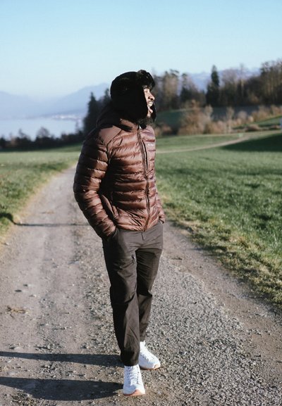 Chaqueta acolchada marrón con capucha con ribete de piel, pantalones negros y zapatillas blancas en un camino de grava con césped verde y montañas al fondo.