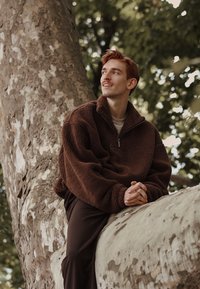 Brauner Fleece-Quarter-Zip-Pullover mit strukturiertem Finish, kombiniert mit dunklen Hosen. Die Person sitzt auf einem großen Baumast.