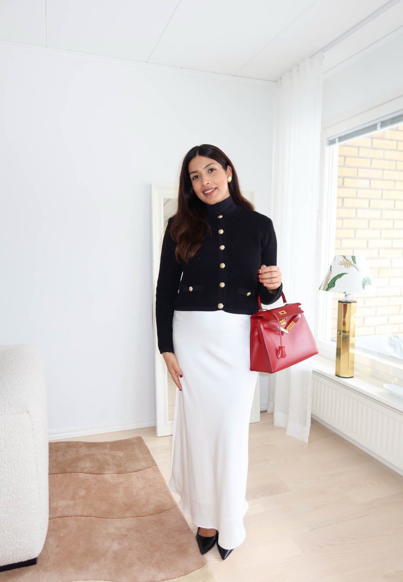 Femme debout à l'intérieur, portant un cardigan noir boutonné, une jupe blanche, des chaussures noires pointues, tenant un sac à main rouge.