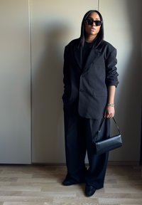 Blazer noir surdimensionné associé à un pantalon large noir, un haut noir simple, un sac à main noir et des chaussures noires épaisses. Tissu texturé.