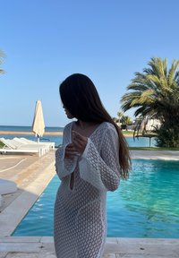 Abito coprente bianco all'uncinetto con maniche lunghe e scollatura a chiave, indossato davanti a uno sfondo di piscina e palme.