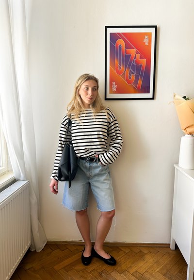 Modelo vistiendo una camisa de rayas en blanco y negro de manga larga, pantalones cortos de mezclilla azul claro y zapatos planos negros, sosteniendo un bolso negro.