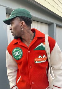 Casaco varsity vermelho e branco com patches verdes, incluindo montanhas e texto, combinado com um boné verde e um brinco em forma de cruz. Saco tote neutro.