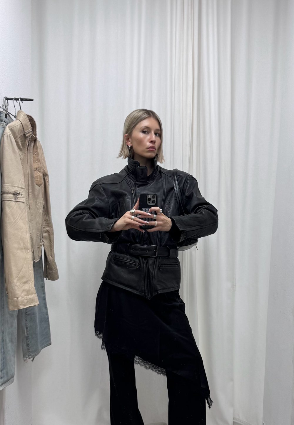 Femme aux cheveux blonds courts portant une veste en cuir noire et un pantalon noir prend un selfie dans un miroir dans une pièce avec des vestes beige et bleues suspendues.