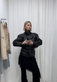 Femme aux cheveux blonds courts portant une veste en cuir noire et un pantalon noir prend un selfie dans un miroir dans une pièce avec des vestes beige et bleues suspendues.