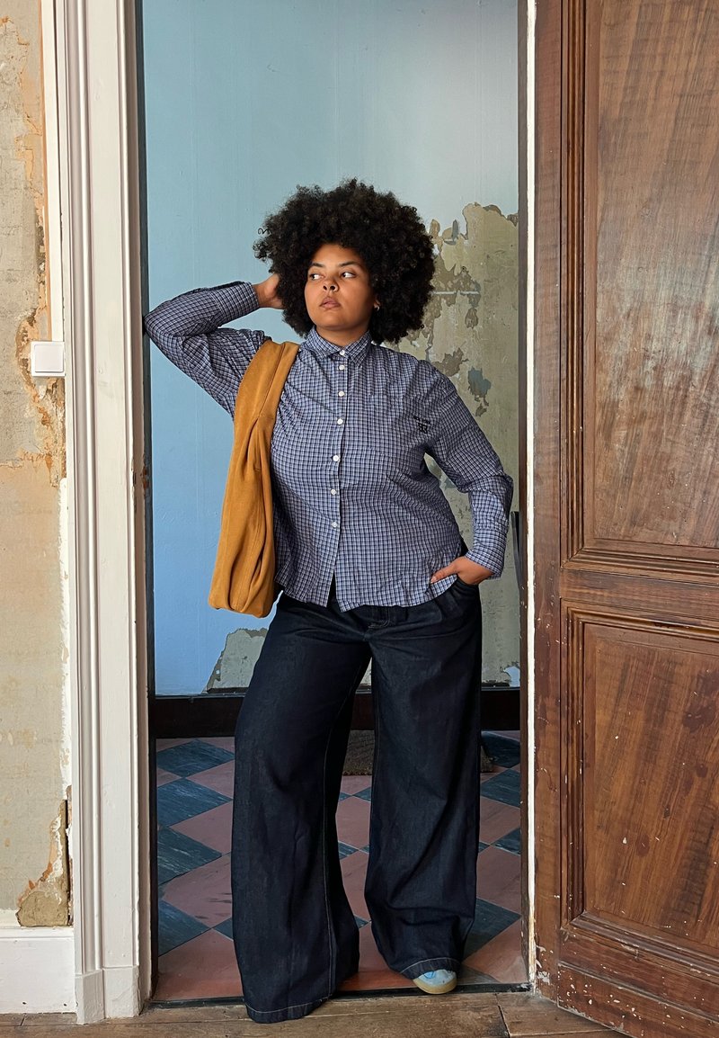 Modelo vestindo uma camisa de botão azul xadrez e jeans largos escuros, com um suéter laranja pendurado sobre um ombro, diante de uma parede texturizada.