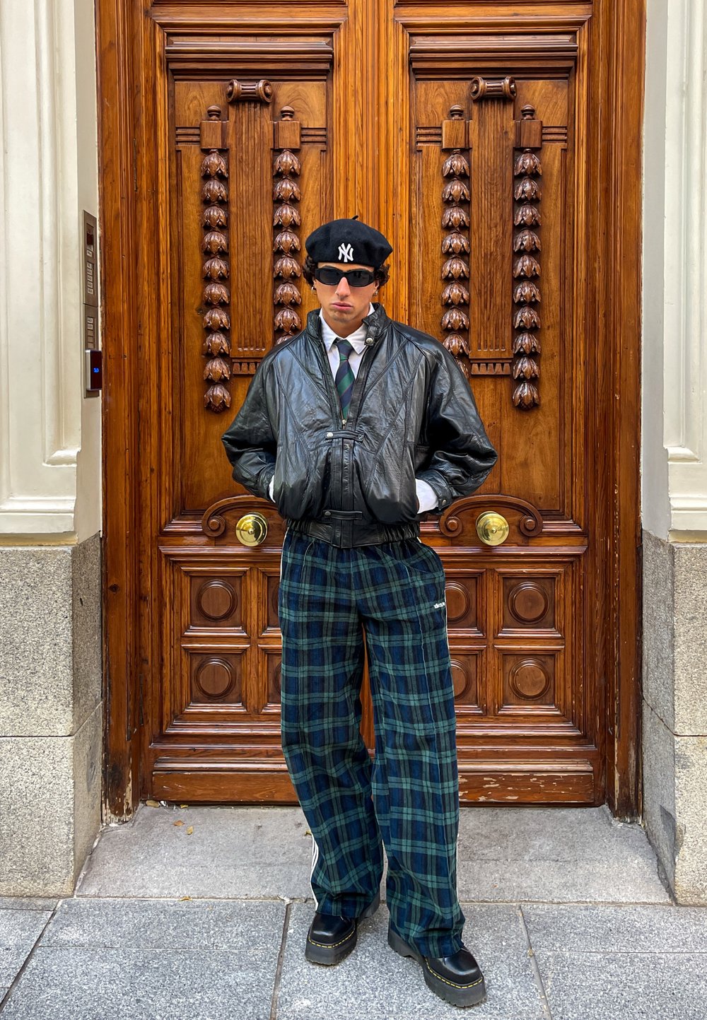 Chaqueta bomber de cuero negro sobre una camisa blanca y corbata a rayas; pantalones de cuadros verdes de pierna ancha y zuecos negros; de pie frente a una puerta de madera.