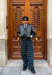 Chaqueta bomber de cuero negro sobre una camisa blanca y corbata a rayas; pantalones de cuadros verdes de pierna ancha y zuecos negros; de pie frente a una puerta de madera.