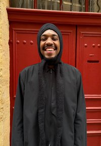 Svart hettejakke med glidelås, sammen med et teksturert svart skjerf og hette, mot en rød dør i bakgrunnen. Stoffet ser mykt og varmt ut.