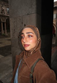 Braune Wildlederjacke mit großem Kragen, kombiniert mit einem gemusterten braun-beigen Schal. Die Person hat helle Haut und blaue Augen.