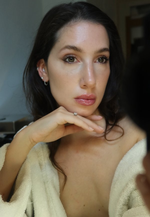 Femme aux cheveux foncés et à la peau nette, reposant le menton sur la main, portant un peignoir doux et plusieurs boucles d'oreilles, regardant pensivement sur le côté.