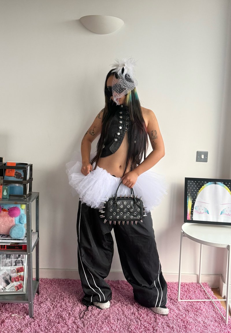 Personne portant un haut noir sans manches, une jupe tutu blanche, un pantalon noir ample, un masque facial avec un accessoire plume blanche, tenant un sac à main noir clouté.