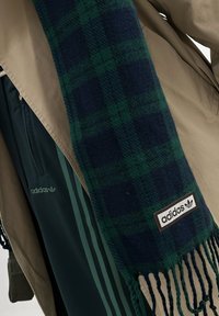 Marineblå og grønn rutete skjerf med frynser og en brun adidas-logo lap, brukt sammen med en beige jakke og grønne treningsbukser.