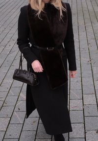 Manteau noir avec une écharpe en fausse fourrure marron, cintré à la taille avec une ceinture noire. Sac à main sombre à motif crocodile avec des anses courtes.