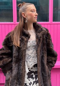 Casaco de peluche castanho com lapelas largas e um design texturizado, sobreposto a uma blusa de renda transparente e um vestido com padrão de vaca preto e branco.