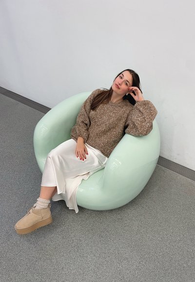 Silla curva, brillante y de color verde menta en forma de haba con una textura suave. Una persona sentada lleva un suéter marrón de punto y una falda blanca. También son visibles unos zapatos beige.