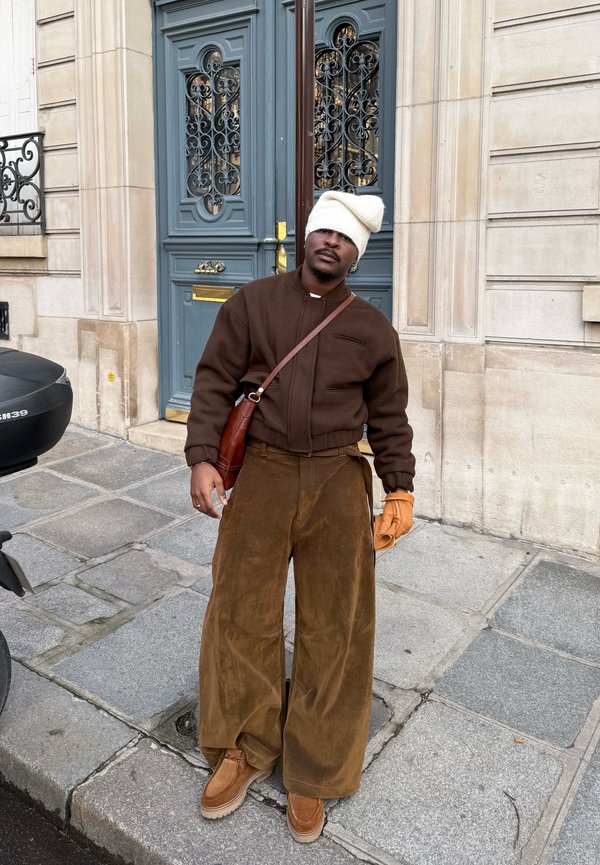 Man draagt een bruin jasje, broek met wijde pijpen, beige laarzen, oranje handschoen en een slordige witte muts, staande op een stadsstoep voor een blauwe deur.