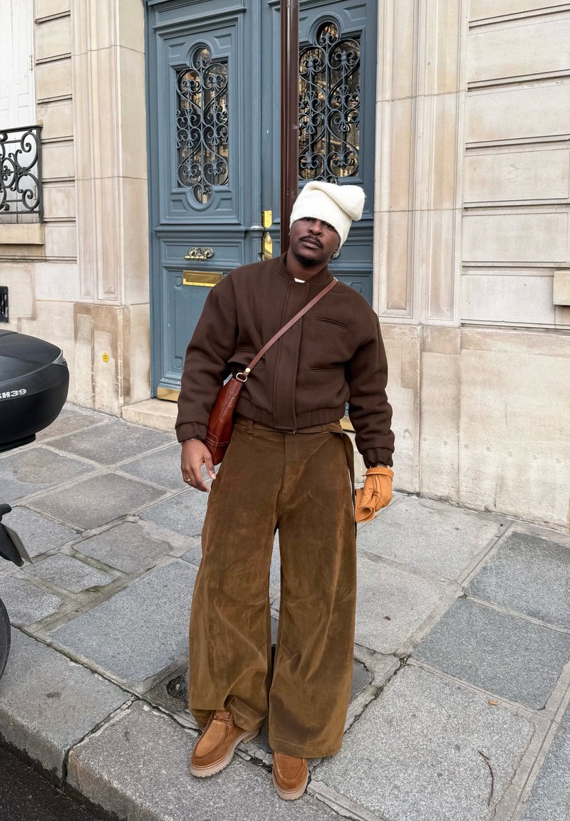 Άνδρας που φορά καφέ μπουφάν, φαρδιά παντελόνια, καφέ μπότες, πορτοκαλί γάντι και χαλαρό λευκό σκουφάκι, στέκεται σε πεζοδρόμιο της πόλης μπροστά από μπλε πόρτα.