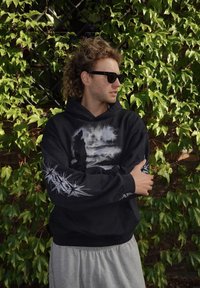 Black hoodie with a nature-inspired graphic print, featuring a figure and abstract elements. Accents on sleeves resemble star patterns. Gray sweatpants.