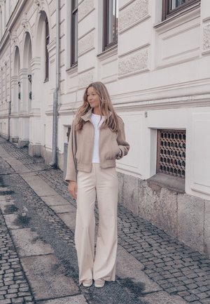 Mujer con cabello largo que lleva chaqueta beige y pantalones de pierna ancha, de pie en una calle empedrada junto a un edificio blanco ornamentado.