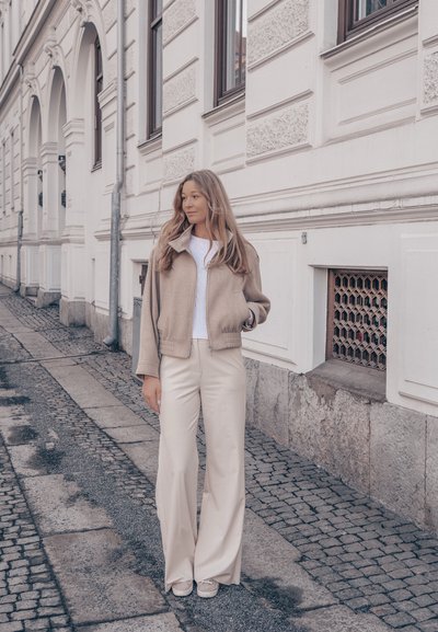 Mujer con cabello largo que lleva chaqueta beige y pantalones de pierna ancha, de pie en una calle empedrada junto a un edificio blanco ornamentado.