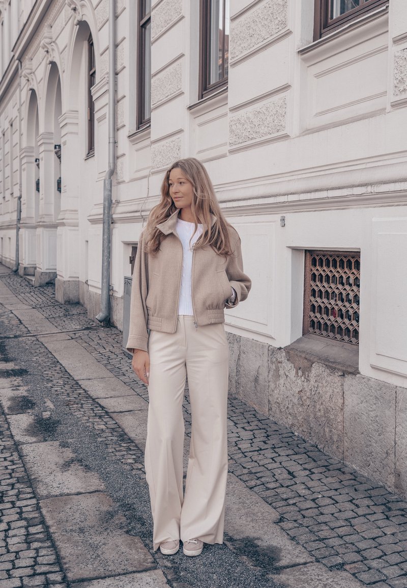 Femme aux longs cheveux portant une veste beige et un pantalon à jambes larges, debout sur une rue pavée à côté d'un bâtiment blanc orné.