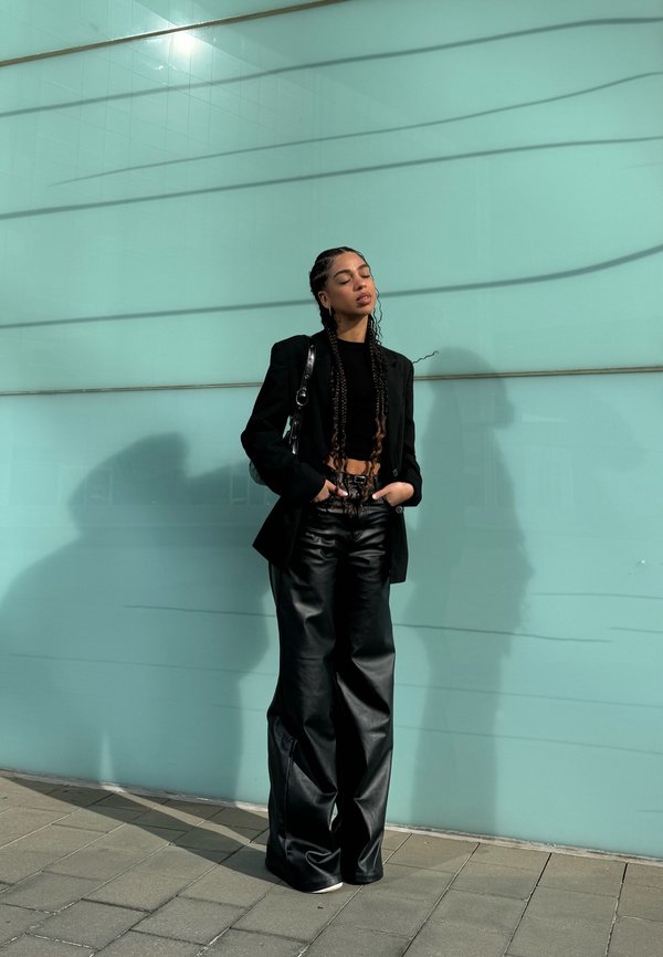 Sorte læder bukser med bred ben, cropped sort top og tætsiddende sort blazer. Modellen står mod en lys teal væg og kaster en skygge.