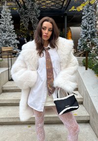 Femme portant un manteau en fausse fourrure blanc, une chemise blanche avec une cravate à motif, des collants en dentelle, tenant un sac à main noir et blanc, debout près d'arbres artificiels enneigés en plein air.