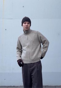 Pull beige en tricot avec un col montant et une fermeture éclair, motif texturé, associé à des gants brun foncé et un pantalon gris ample.
