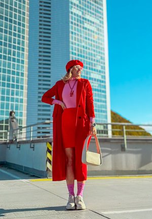 Abrigo largo rojo sobre una blusa rosa, falda roja, zapatos blancos, calcetines rosas con patrones y un bolso crema. Fondo de un entorno urbano moderno.
